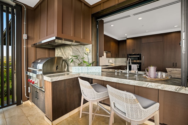 kitchen featuring a breakfast bar area, light stone counters, kitchen peninsula, wall chimney range hood, and backsplash