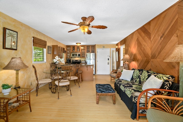 living room featuring wooden walls, ceiling fan, a textured ceiling, and light wood-type flooring