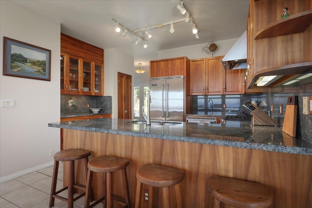 kitchen with backsplash, exhaust hood, kitchen peninsula, light tile patterned flooring, and stainless steel built in refrigerator