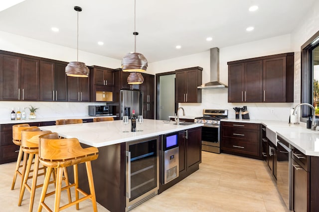 kitchen with appliances with stainless steel finishes, wall chimney exhaust hood, wine cooler, hanging light fixtures, and a center island with sink