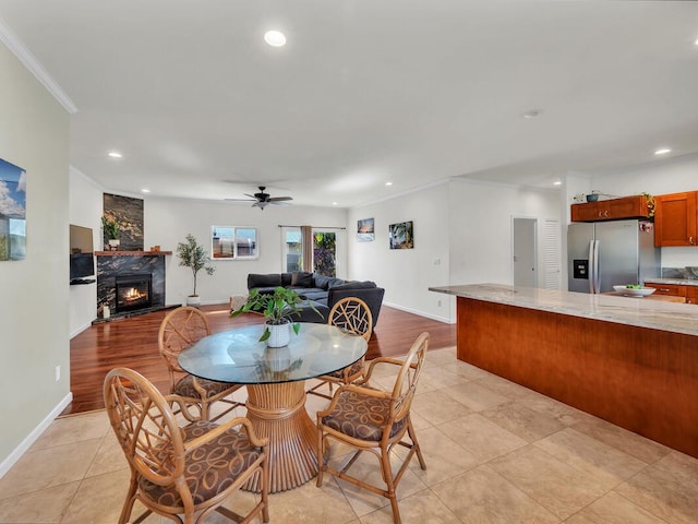 dining room featuring light tile patterned flooring, ceiling fan, crown molding, and a stone fireplace