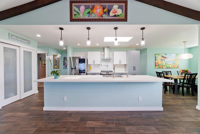 kitchen featuring wall chimney exhaust hood, white cabinetry, a center island with sink, pendant lighting, and stainless steel appliances