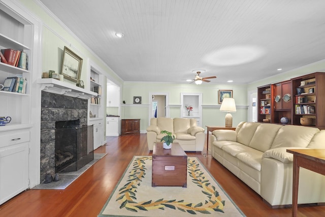 living room featuring ceiling fan, crown molding, built in features, dark hardwood / wood-style floors, and a stone fireplace