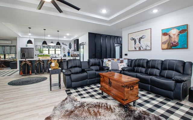 living room with light wood-type flooring, a wall mounted air conditioner, ceiling fan, and a tray ceiling