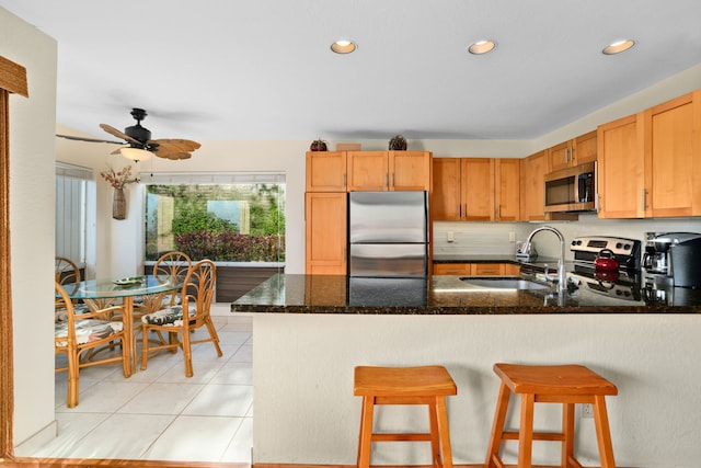 kitchen with kitchen peninsula, stainless steel appliances, ceiling fan, sink, and dark stone countertops