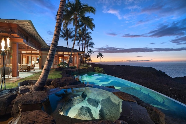 pool at dusk featuring an in ground hot tub, a patio, and a water view