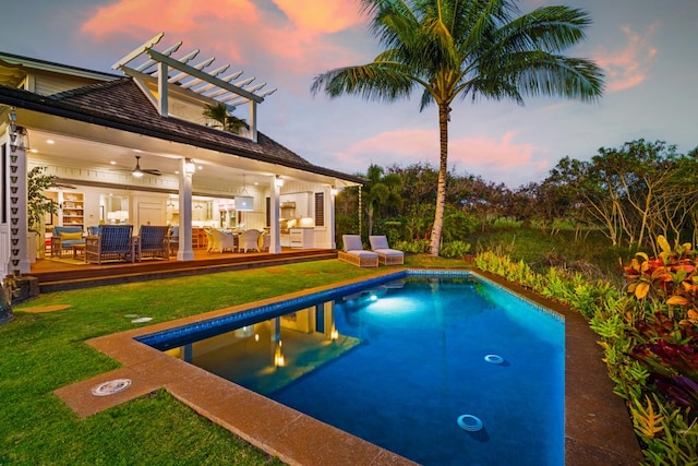 pool at dusk featuring a wooden deck, ceiling fan, outdoor lounge area, and a lawn