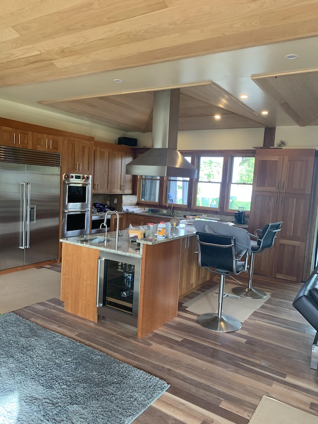 kitchen with light stone countertops, appliances with stainless steel finishes, island exhaust hood, wine cooler, and a center island with sink