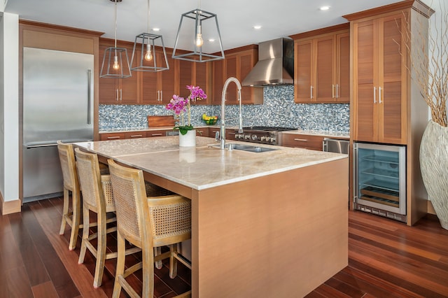 kitchen with pendant lighting, wine cooler, a center island with sink, built in fridge, and wall chimney exhaust hood