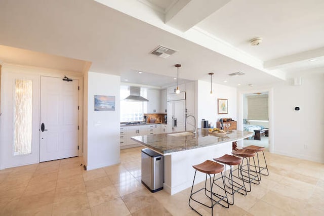 kitchen featuring visible vents, a breakfast bar, wall chimney range hood, white cabinetry, and built in fridge