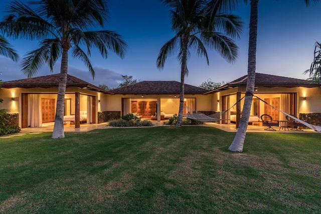 back house at dusk featuring a lawn and a patio area