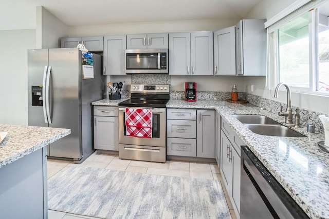 kitchen with light tile patterned floors, stainless steel appliances, gray cabinets, a sink, and light stone countertops