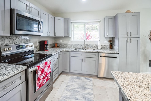 kitchen with light tile patterned floors, gray cabinets, appliances with stainless steel finishes, a sink, and light stone countertops