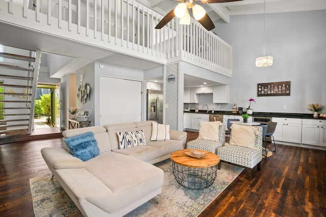 living area with dark wood finished floors, beam ceiling, ceiling fan with notable chandelier, and a towering ceiling