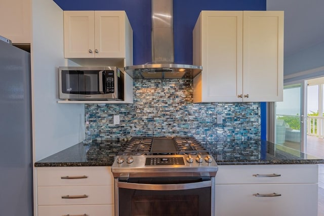 kitchen with wall chimney exhaust hood, dark stone countertops, stainless steel appliances, decorative backsplash, and white cabinets