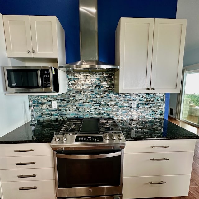 kitchen featuring stainless steel appliances, dark stone countertops, wall chimney range hood, and white cabinets