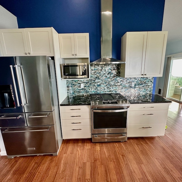 kitchen with wall chimney exhaust hood, light wood-type flooring, dark stone countertops, stainless steel appliances, and white cabinets