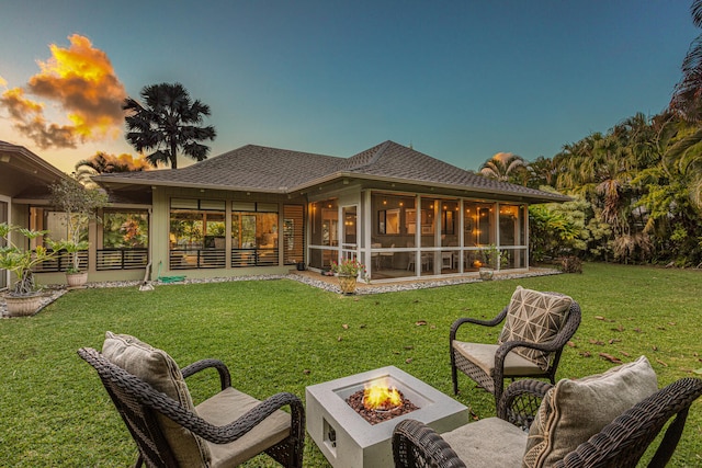 back of house at dusk with an outdoor fire pit, a sunroom, roof with shingles, and a yard