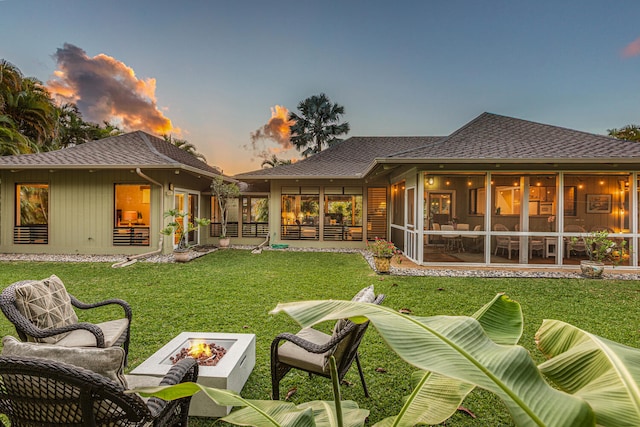 back of property at dusk featuring a sunroom, roof with shingles, a yard, and a fire pit