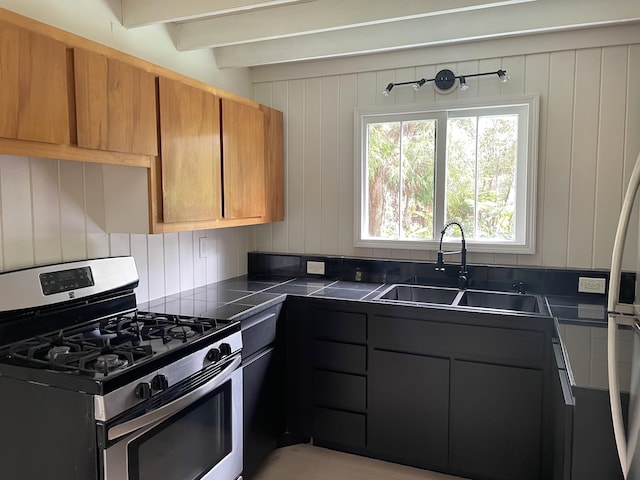 kitchen with tile countertops, a sink, brown cabinetry, stainless steel gas stove, and beamed ceiling