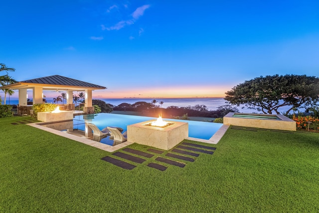 pool at dusk featuring a gazebo, a water view, an outdoor fire pit, and a lawn