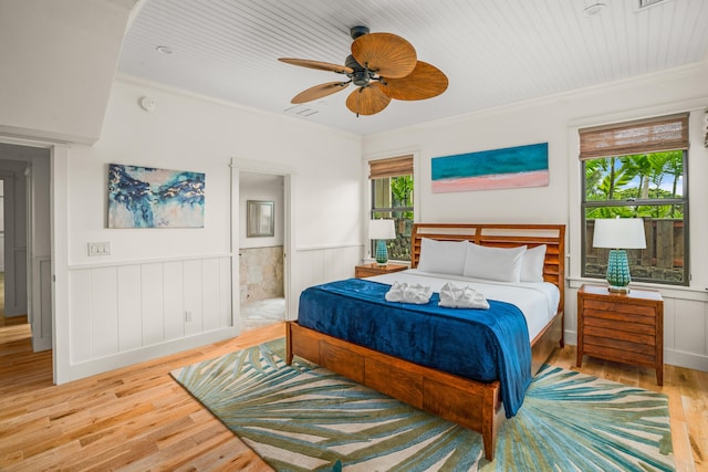 bedroom featuring wainscoting, ceiling fan, wood-type flooring, ensuite bathroom, and crown molding