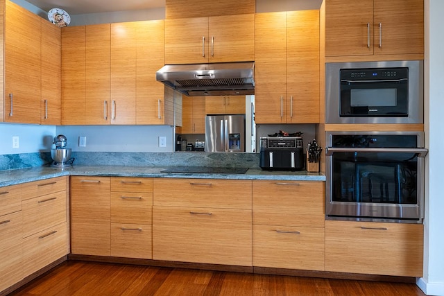 kitchen featuring stone counters, dark wood-type flooring, under cabinet range hood, black electric cooktop, and stainless steel fridge