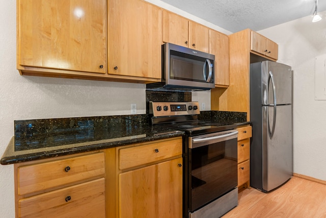 kitchen featuring a textured ceiling, baseboards, appliances with stainless steel finishes, light wood finished floors, and dark stone countertops