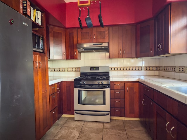 kitchen featuring tile patterned flooring, under cabinet range hood, appliances with stainless steel finishes, tile counters, and tasteful backsplash