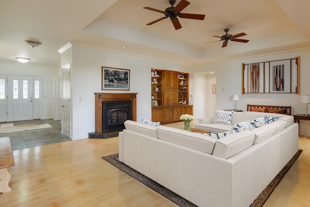 living room with wood finished floors, ornamental molding, a fireplace with raised hearth, ceiling fan, and a tray ceiling