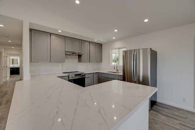 kitchen with light stone countertops, under cabinet range hood, gray cabinets, appliances with stainless steel finishes, and a sink