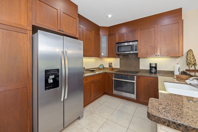 kitchen with a sink, dark stone countertops, appliances with stainless steel finishes, brown cabinetry, and light tile patterned floors