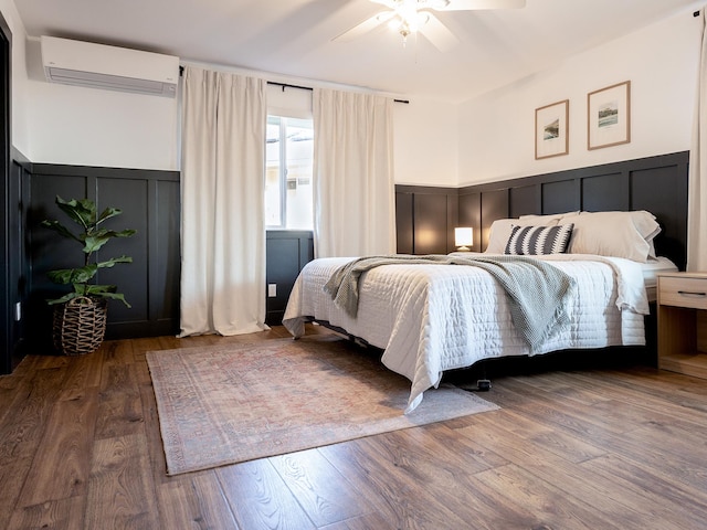 bedroom featuring wood finished floors, ceiling fan, a decorative wall, a wall mounted air conditioner, and wainscoting