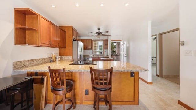 kitchen with freestanding refrigerator, a peninsula, brown cabinetry, light stone counters, and a ceiling fan