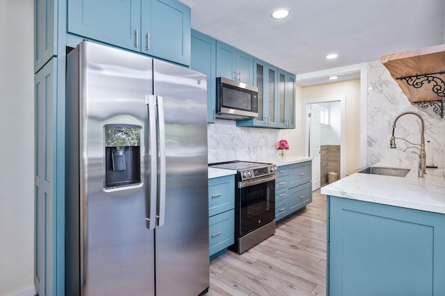kitchen featuring appliances with stainless steel finishes, a sink, blue cabinetry, light wood-type flooring, and glass insert cabinets