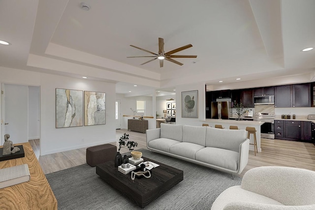 living area with light wood-type flooring, recessed lighting, ceiling fan, and a tray ceiling