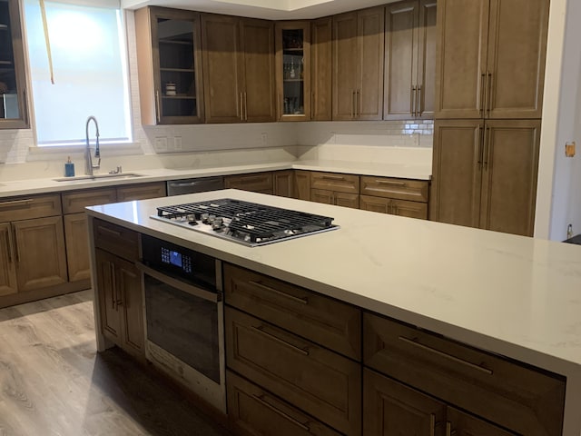 kitchen with a sink, light wood-style floors, stainless steel appliances, backsplash, and glass insert cabinets