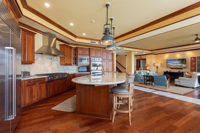 kitchen with wall chimney exhaust hood, built in appliances, brown cabinets, a ceiling fan, and dark wood-style floors
