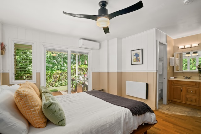 bedroom with an AC wall unit, light wood-type flooring, radiator heating unit, ensuite bathroom, and a ceiling fan