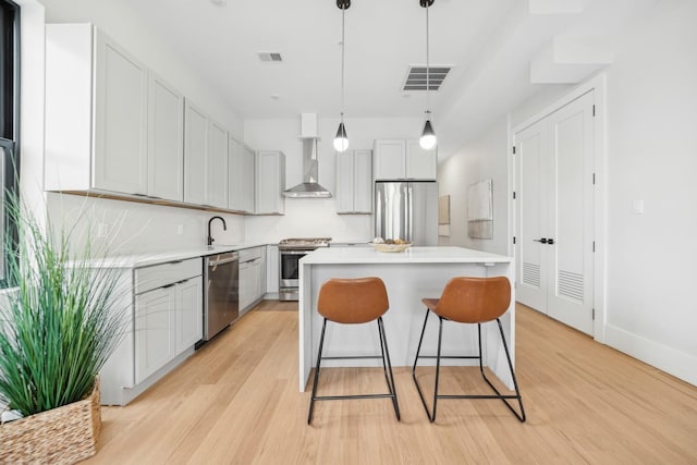 kitchen with wall chimney range hood, light hardwood / wood-style flooring, pendant lighting, a kitchen island, and appliances with stainless steel finishes