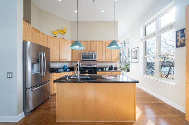 kitchen with light brown cabinets, a kitchen island with sink, appliances with stainless steel finishes, dark countertops, and tasteful backsplash