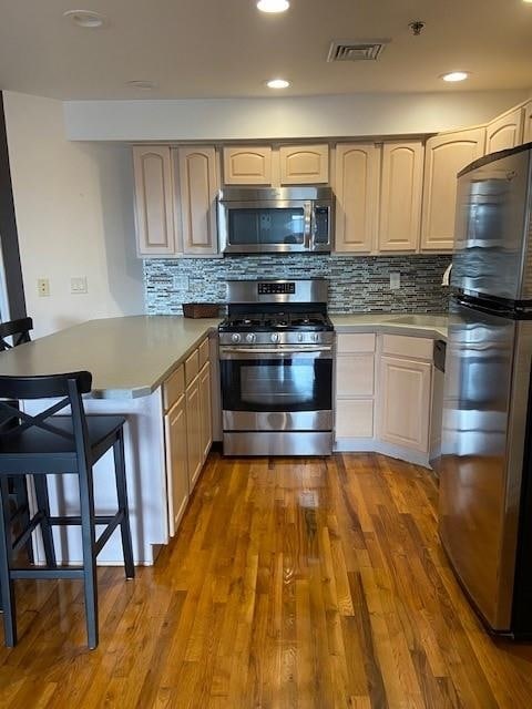 kitchen featuring light brown cabinetry, decorative backsplash, hardwood / wood-style flooring, kitchen peninsula, and stainless steel appliances