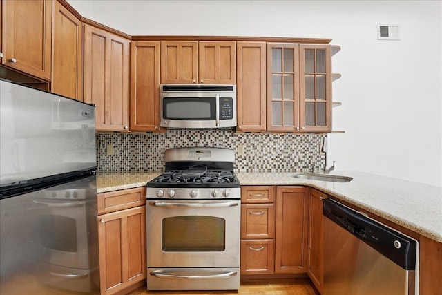 kitchen featuring appliances with stainless steel finishes, decorative backsplash, brown cabinetry, and glass insert cabinets
