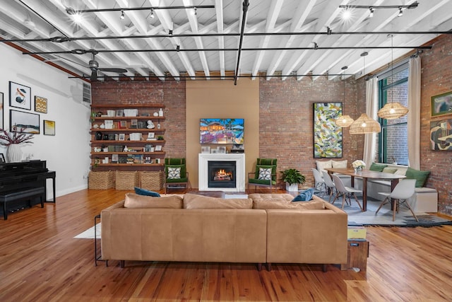 living room featuring a warm lit fireplace, wood finished floors, brick wall, rail lighting, and baseboards