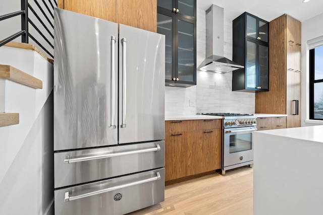 kitchen with tasteful backsplash, wall chimney exhaust hood, appliances with stainless steel finishes, light countertops, and light wood-style floors