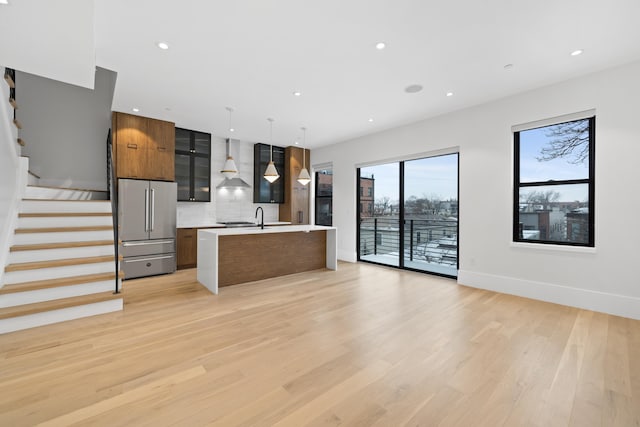 kitchen featuring high end refrigerator, a kitchen island with sink, light countertops, light wood-type flooring, and wall chimney range hood