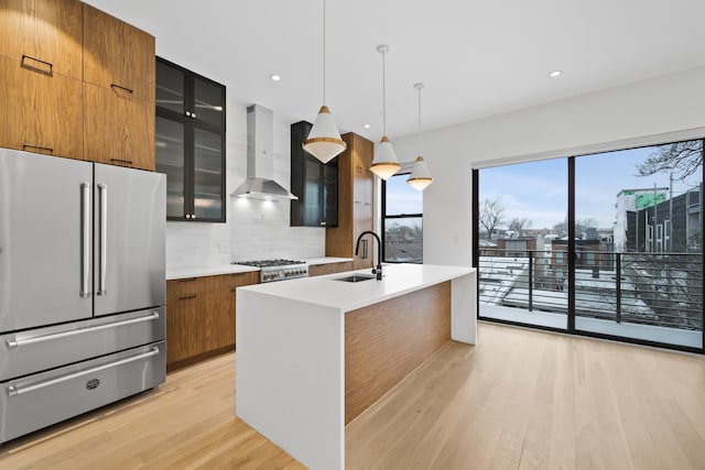 kitchen featuring high end refrigerator, brown cabinetry, a sink, modern cabinets, and wall chimney exhaust hood