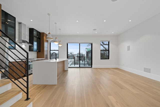 kitchen with light wood-style floors, a kitchen island with sink, light countertops, and wall chimney range hood