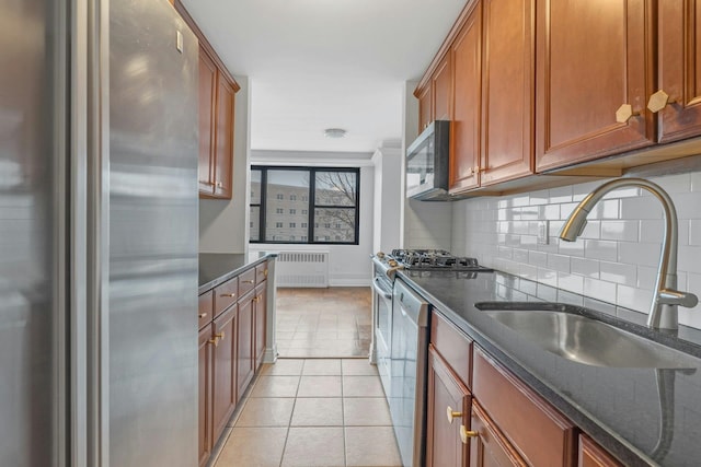 kitchen featuring stainless steel appliances, light tile patterned floors, dark stone counters, radiator heating unit, and sink