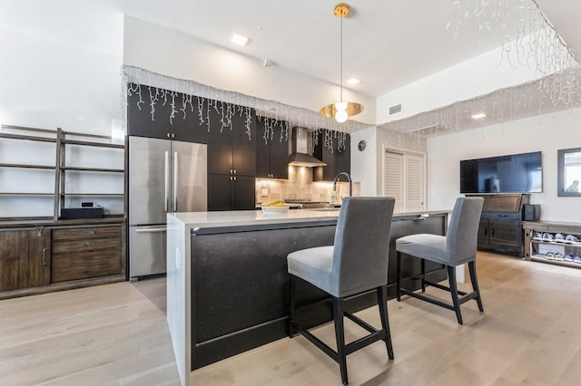 kitchen featuring visible vents, wall chimney range hood, light countertops, light wood-style flooring, and stainless steel refrigerator
